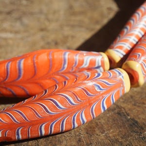 May include: Close-up of several orange glass beads with blue and white swirl patterns. The beads have yellow ends and are arranged on a wooden surface. The beads are elongated and cylindrical in shape.