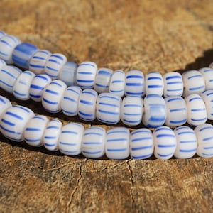 Pode incluir: Close-up de três fios de contas brancas com listras horizontais azuis. As contas são cilíndricas e dispostas em um padrão curvo. O fundo é uma superfície de madeira marrom texturizada.