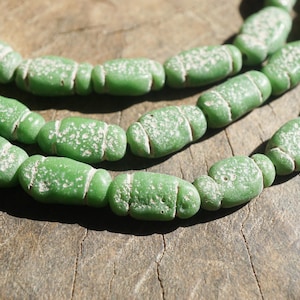 Può includere: Primo piano di una collana di perline di vetro verde. Le perline sono di forma ovale con una superficie strutturata e dettagli bianchi. Le perline sono disposte su una superficie di legno.