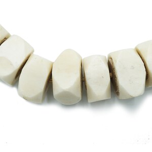 May include: Close-up of a necklace featuring large, off-white, geometric beads. The beads have a faceted design and are strung together with a gold-colored cord. The background is plain white.