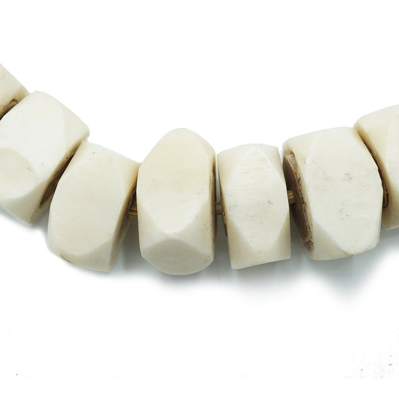 May include: Close-up of a necklace featuring large, off-white, geometric beads. The beads have a faceted design and are strung together with a gold-colored cord. The background is plain white.