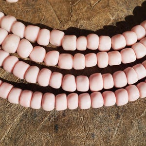 May include: Close-up of several strands of light pink beads. The beads are a soft, matte finish and are strung together in a curved arrangement. The background is a textured, brown surface, providing a contrast to the delicate color of the beads.