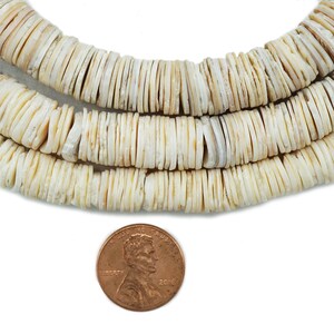 Op de afbeelding: Een streng witte schelp kralen, elke kraal is een platte, ronde schijf. De kralen zijn gerangschikt in een cirkelvormig patroon, waarbij de kralen in het midden van de cirkel iets kleiner zijn dan de kralen aan de buitenkant van de cirkel. Een cent is opgenomen voor de schaal.