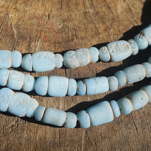 May include: Three strands of light blue, barrel-shaped beads. The beads have a slightly rough texture and are strung together. The background is a weathered, brown wooden surface. The beads are a soft, muted blue color.