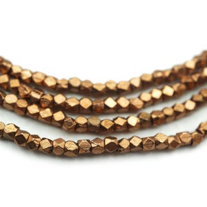 Puede incluir: Primer plano de varios hilos de cuentas facetadas de color cobre. Las cuentas tienen una forma geométrica y un acabado metálico. Las cuentas están ensartadas, creando un componente de collar o pulsera.