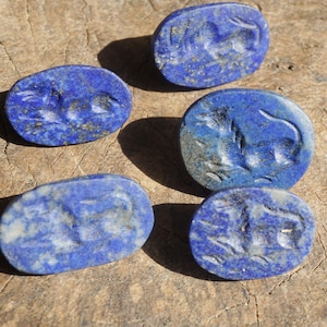 May include: Five oval-shaped, deep blue lapis lazuli stones with carved animal figures. The stones are arranged on a rough, wooden surface, with shadows adding depth. The carvings appear to be of a horse or similar animal.