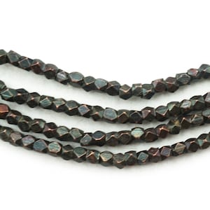 Puede incluir: Primer plano de varios hilos de cuentas facetadas de color bronce oscuro. Las cuentas tienen una forma geométrica y un brillo metálico, con toques de azul y verde. Las cuentas están ensartadas, para joyería.