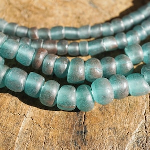 Pode incluir: Close-up de vários fios de contas de vidro turquesa. As contas são cilíndricas e têm uma forma ligeiramente irregular, com uma qualidade translúcida. As contas são enfiadas juntas, criando um colar ou uma pulseira. O fundo é uma superfície de madeira texturizada.