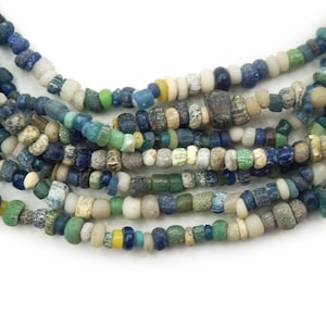 Può includere: Fili di perline di vetro antiche e colorate in tonalità blu, verde, bianco e giallo. Le perline variano per dimensioni e forma, creando un aspetto strutturato e unico. Le perline sono infilate, creando una collana o un braccialetto.