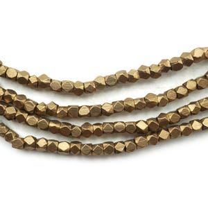 Puede incluir: Primer plano de varios hilos de cuentas facetadas de color bronce. Las cuentas tienen una forma geométrica y están ensartadas, creando una apariencia texturizada y detallada. Las cuentas están sobre un fondo blanco.