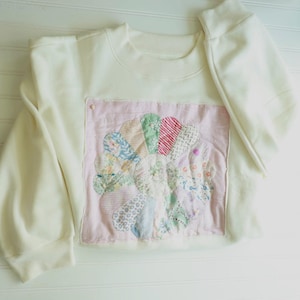 May include: A cream-colored long-sleeve sweatshirt featuring a square patch on the front. The patch is light pink and displays a circular floral design made of various patterned fabric petals in shades of green, blue, red, and white.