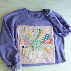 May include: A lavender long-sleeve crewneck sweatshirt featuring a large, square, light pink fabric patch on the front. The patch displays a circular flower design made of various multi-colored patterned fabric petals.