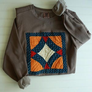 May include: A brown crewneck sweatshirt featuring a square quilted patch on the front. The patch displays a geometric pattern in orange, cream, and dark blue with small white floral details and red accents. The garment has long sleeves with ribbed cuffs.