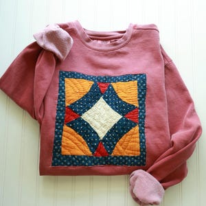 May include: A dusty rose crewneck sweatshirt featuring a large, square, quilted patch on the front. The patch displays a geometric pattern in cream, orange, red, and dark blue with a small floral print on the dark blue fabric sections.
