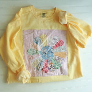 May include: A pale yellow long-sleeve sweatshirt featuring a large square patch on the front. The patch displays a multi-colored floral design made of various patterned fabric petals stitched onto a light pink background fabric.
