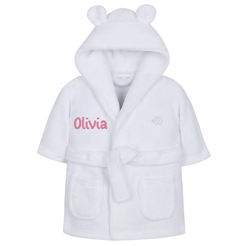 May include: White hooded bathrobe with a pink embroidered name "Olivia" and a small elephant logo. The robe has a tie belt and pockets.