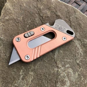 May include: A copper-coloured utility knife with a retractable blade, featuring a grey metal top section engraved with the text T&P. The tool includes a sliding mechanism, three visible screws, and a cutout handle design with a bottle opener.