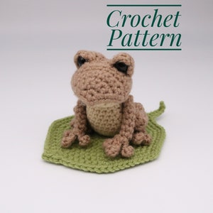 May include: A crocheted brown frog sitting on a green crocheted leaf. The frog has black eyes and a light brown belly. The text "Crochet Pattern" is written in green above the frog.
