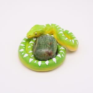 May include: A handmade, green and yellow snake figurine coiled around a green and brown stone. The snake has white markings along its body and is set against a white background. The figurine is likely made of ceramic or a similar material.