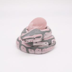 Puede incluir: Figurita de serpiente de cerámica enroscada, rosa y gris. El cuerpo de la serpiente tiene marcas grises sobre una base rosa. La cabeza presenta un pequeño ojo plateado. Un objeto rosa en forma de huevo descansa sobre el cuerpo enroscado.