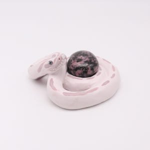 May include: A white ceramic snake figurine with pink markings, coiled around a dark, speckled sphere. The snake has a detailed head with a small, round eye. The overall design is artistic and decorative.