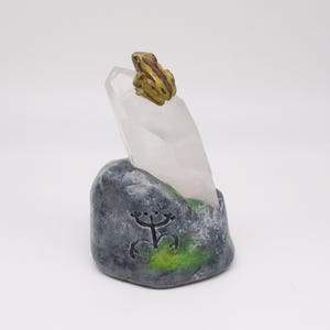 May include: A decorative sculpture featuring a clear quartz crystal emerging from a gray, stone-like base with green accents. A small, detailed frog figurine sits atop the crystal. The base has a carved symbol.