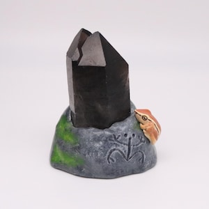 May include: A dark, smoky quartz crystal stands on a gray, stone-like base with green moss accents. A small, orange and brown frog figurine is perched on the side. The base features a carved symbol.
