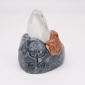 May include: A clear quartz crystal sits atop a dark gray stone base with a carved symbol. A small, brown frog figurine is perched on the side. The base has a rough texture, and the crystal is pointed.