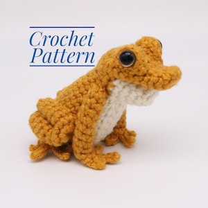 Puede incluir: Patrón de crochet para una pequeña rana amarilla con vientre blanco. La rana está sentada sobre una superficie blanca.