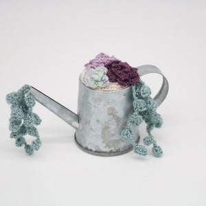 May include: A small metal watering can with a crocheted succulent plant inside. The plant is a mix of purple, pink, and green.