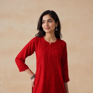 May include: A vibrant red tunic featuring three-quarter length sleeves, a notched round neckline, and intricate tonal embroidery detailing across the chest and hemline. The garment is paired with blue denim jeans, creating a casual ensemble.