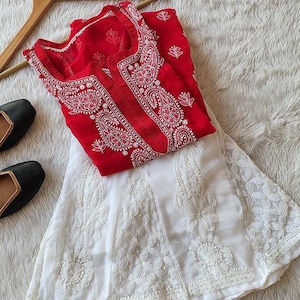 Könnte beinhalten: Ein weißes und rotes besticktes Kleid mit einem weißen Rock und einem roten Oberteil. Der Rock hat ein Blumenmuster und das Oberteil ein Paisley-Muster. Das Kleid liegt auf einer weißen, flauschigen Oberfläche.