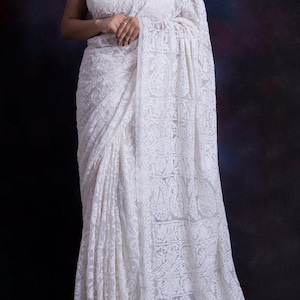 May include: A white saree featuring intricate tone-on-tone floral and paisley embroidery throughout the fabric. The garment includes a sleeveless blouse with a high neckline and a long, draped pallu that extends to the floor with detailed needlework.