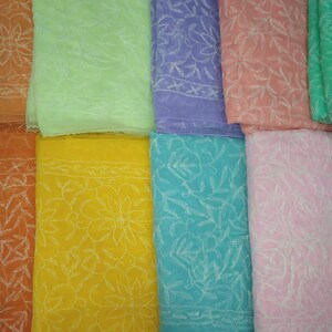 Op de afbeelding: Acht verschillende gekleurde vierkanten van stof met witte geborduurde bloemenpatronen. De kleuren zijn oranje, lichtgroen, paars, roze, geel, turquoise, roze en groen.