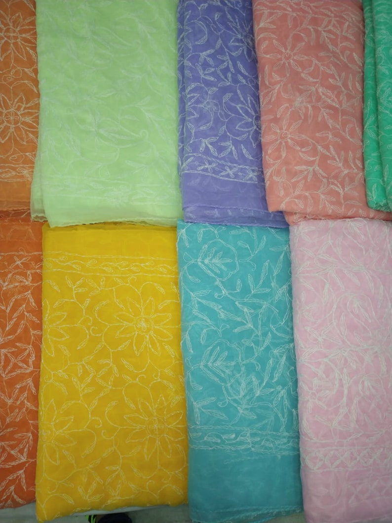 Op de afbeelding: Acht verschillende gekleurde vierkanten van stof met witte geborduurde bloemenpatronen. De kleuren zijn oranje, lichtgroen, paars, roze, geel, turquoise, roze en groen.
