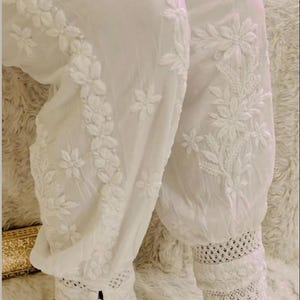 May include: White cotton pants with intricate floral embroidery and lace trim at the bottom. The pants are gathered at the ankles.