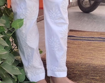 May include: White cotton pants with white embroidered floral designs. The pants are worn with gold embellished flat shoes.