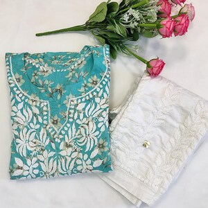 以下が含まれることがあります： 白のパンツとターコイズブルーの花柄刺繍のクルタセット。 クルタは、詳細な白い花柄と丸いネックラインが特徴です。 ピンクのバラと緑のブーケが白い背景に置かれています。