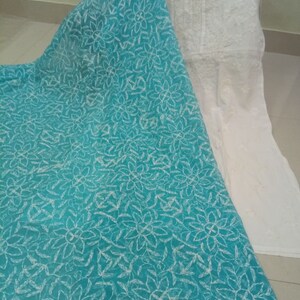 Op de afbeelding: Een witte geborduurde tuniek met een turquoise bloemenpatroon. De tuniek is gemaakt van een transparante stof en heeft een ronde halslijn. De stof is gedrapeerd over een turquoise stof met bloemenpatroon.