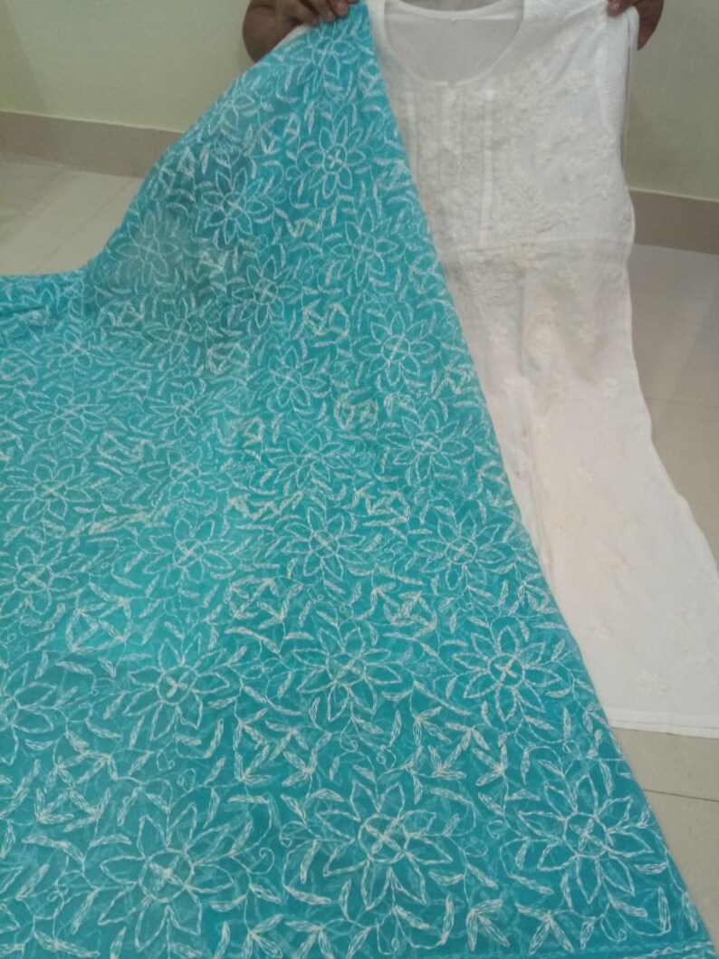 Op de afbeelding: Een witte geborduurde tuniek met een turquoise bloemenpatroon. De tuniek is gemaakt van een transparante stof en heeft een ronde halslijn. De stof is gedrapeerd over een turquoise stof met bloemenpatroon.