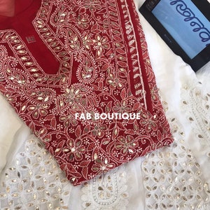Könnte beinhalten: Eine rote und weiße bestickte Kurta mit goldenen Verzierungen. Die Kurta ist aus einem durchsichtigen Stoff gefertigt und hat einen runden Ausschnitt. Der Stoff ist mit komplizierten Blumen- und Paisleymustern verziert. Die Kurta wird mit einer weißen Dupatta mit passenden goldenen Verzierungen kombiniert.