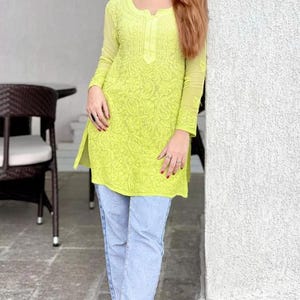 Könnte beinhalten: Eine limettengrüne Tunika mit langen Ärmeln und aufwendiger Stickerei. Die Tunika wird mit hellblauen Jeans kombiniert. Das Outfit wird durch bunte Sandalen ergänzt. Die Tunika hat einen runden Ausschnitt und einen Seitenschlitz.