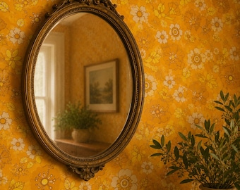 May include: An oval wall mirror with an ornate, dark gold-toned frame featuring decorative scrollwork at the top and bottom. The mirror reflects a room interior with a window, a framed picture on the wall, and a potted plant on a wooden surface.