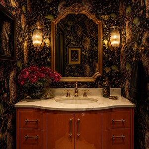 May include: A bathroom vanity with a white marble countertop, an integrated sink, and a glossy orange cabinet featuring gold-toned hardware. Above the vanity is a large gold-framed mirror, two ribbed glass wall sconces, and dark leopard-print wallpaper.