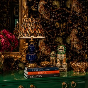 May include: A table lamp with a blue glass base and a leopard print shade sits on a stack of three books titled A Way of Living, Capri Dolce Vita, and Beaton. Beside the books are two ceramic foo dog figurines and a gold decorative bowl.