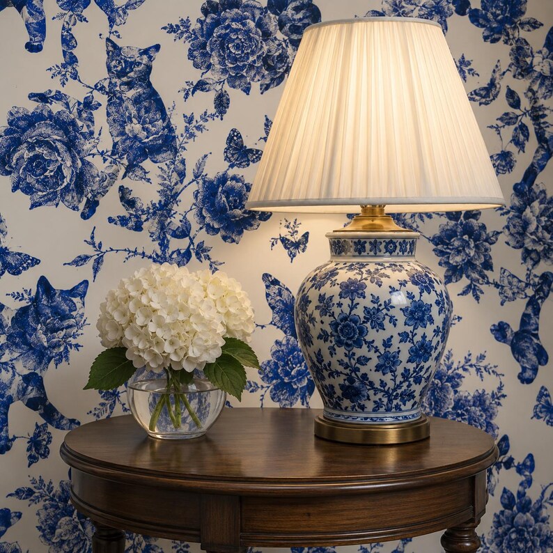 Puede incluir: Una l&aacute;mpara de mesa con una pantalla plisada blanca y una base de cer&aacute;mica con estampado floral azul y blanco. La l&aacute;mpara est&aacute; sobre una mesa de madera oscura, con un peque&ntilde;o cuenco de cristal con flores blancas. Fondo de papel pintado floral azul y blanco.