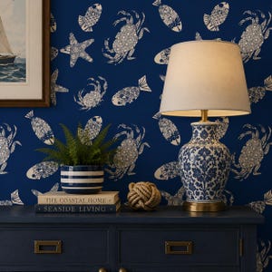 May include: A blue and white ceramic table lamp with a cream-colored lampshade sits on a navy blue dresser. The lamp has a floral design. A small potted plant, books, and a decorative rope knot are also on the dresser. The wallpaper has a nautical theme.