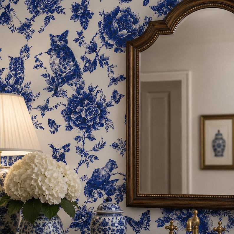 Puede incluir: Una habitaci&oacute;n con papel pintado floral azul y blanco con motivos de gatos y mariposas. Un espejo con marco dorado refleja una puerta y una impresi&oacute;n enmarcada. Una l&aacute;mpara con pantalla blanca y un jarr&oacute;n con flores blancas est&aacute;n sobre una mesa.