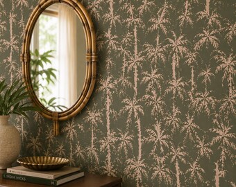 May include: An oval mirror with a bamboo-style frame hangs on a wall covered in olive green wallpaper featuring a repeating light pink palm tree pattern. A stack of books and a small bowl sit on a wooden surface below the mirror.
