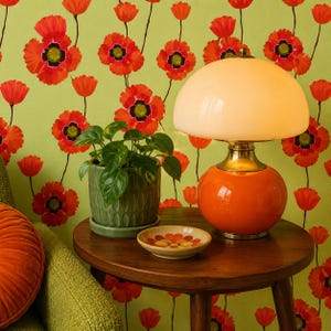 Puede incluir: Una l&aacute;mpara de mesa naranja de estilo retro con una pantalla color crema se encuentra sobre una mesa de madera oscura. Una peque&ntilde;a maceta verde y un plato decorativo con un dise&ntilde;o floral tambi&eacute;n est&aacute;n sobre la mesa. El fondo presenta una pared verde con un patr&oacute;n de amapolas.